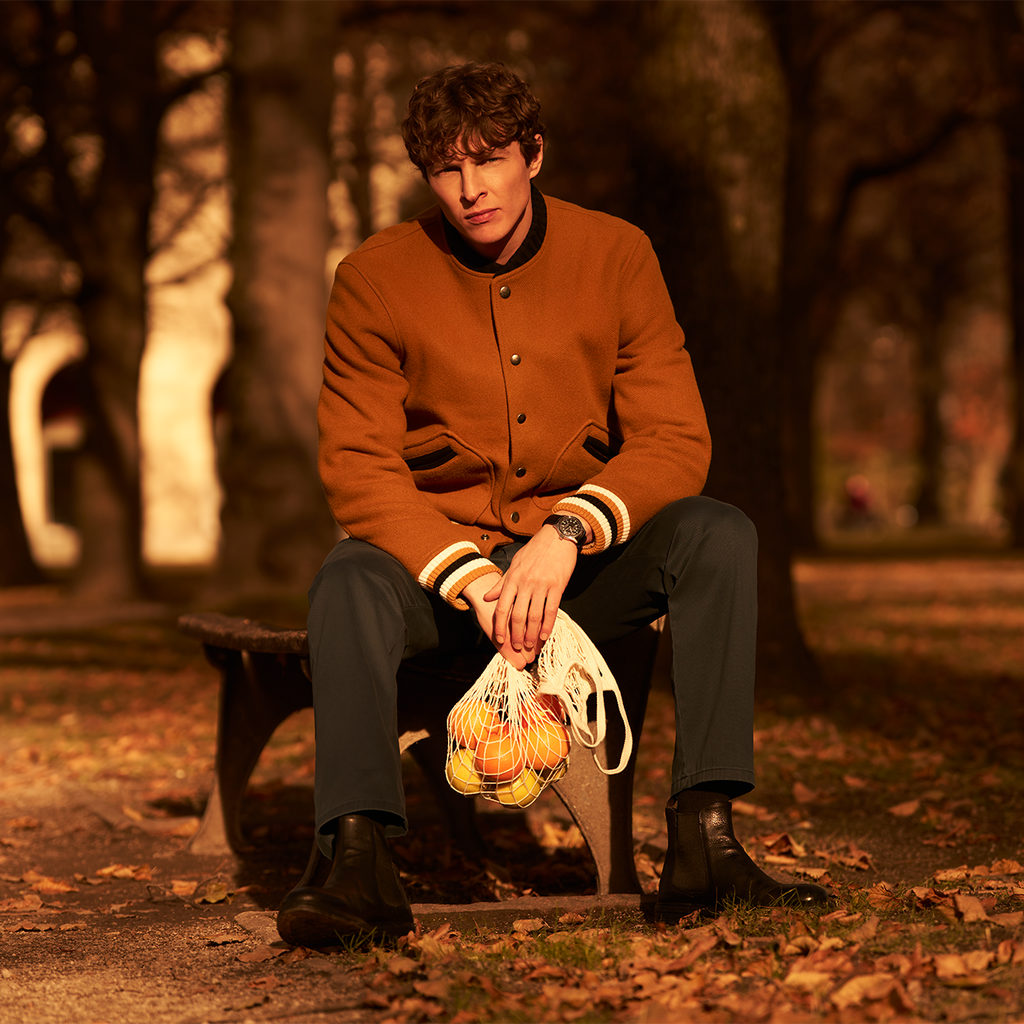 Person sitting on a bench in a park holding a bag of groceries, surrounded by trees and fallen leaves.